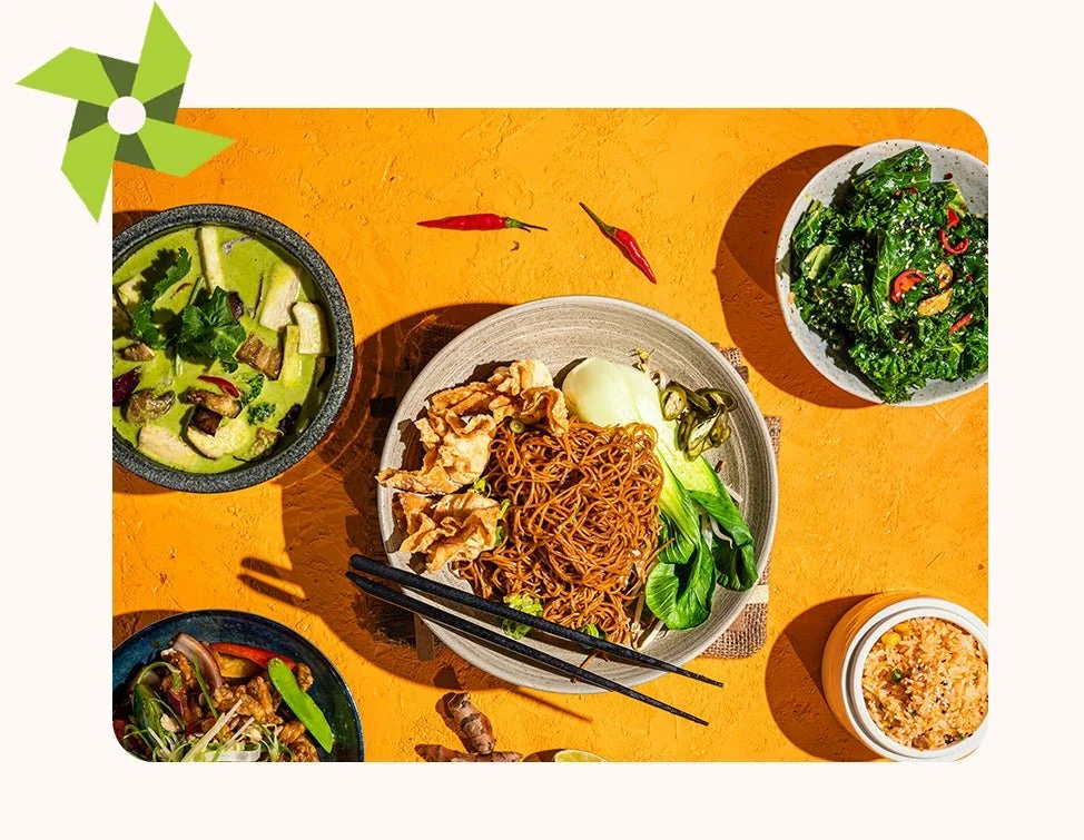 An assortment of Asian dishes on an orange table, including a plate of noodles with vegetables, bowls of salad, a soup, and a container of fried rice, with red chili peppers as garnish.