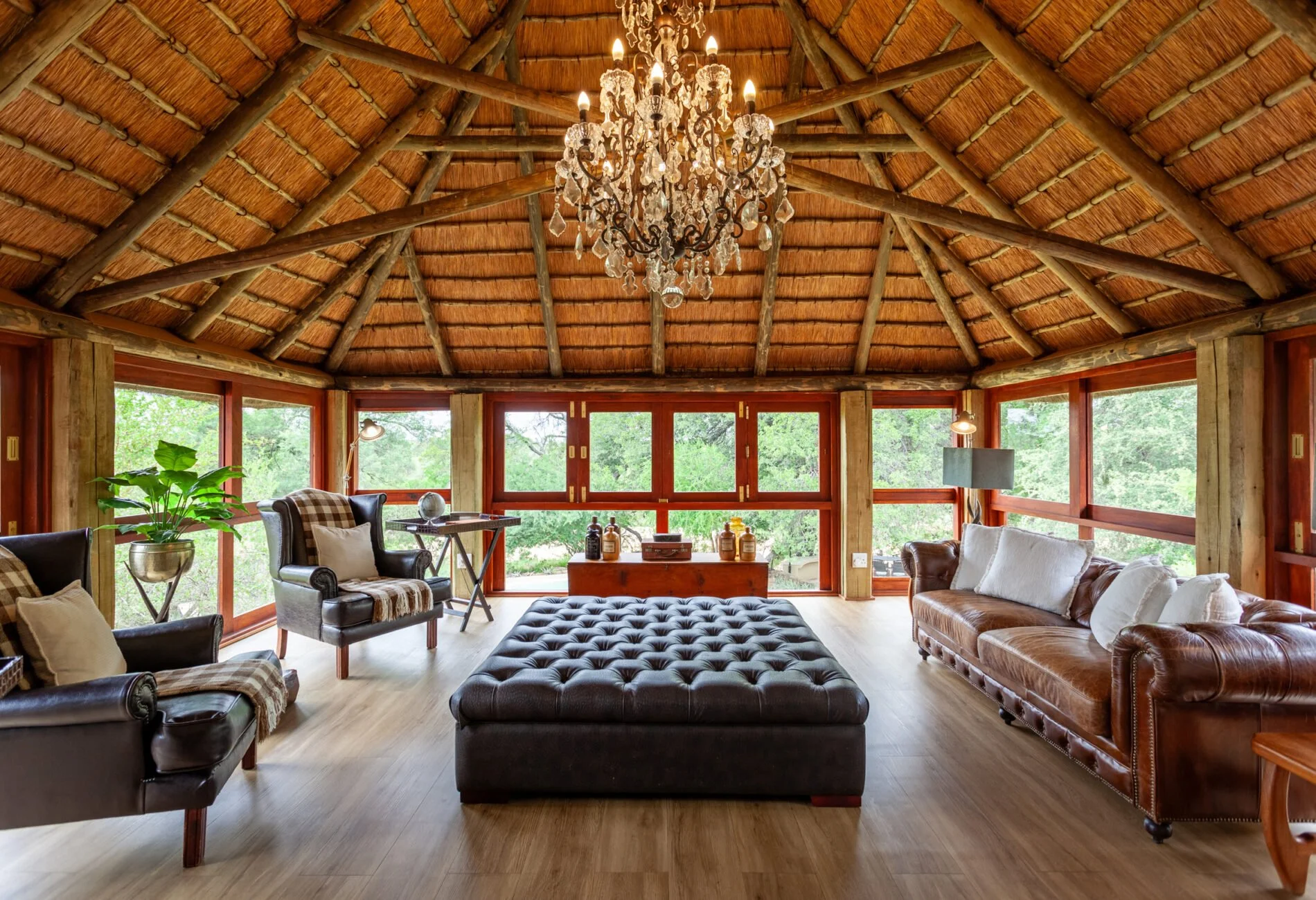Rustic living room with large windows, wooden walls and ceiling, leather and fabric armchairs, a tufted ottoman, a chandelier, and various decorative items.