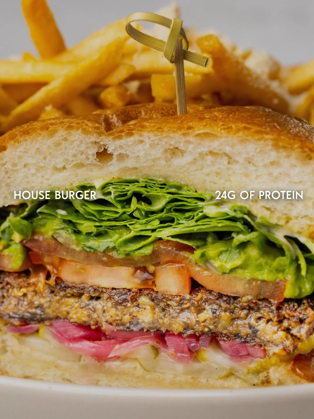 Close-up of a house burger with lettuce, tomato, cheese, and a patty, cut open to show fillings, with French fries in the background.