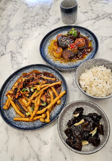 Four dishes on a marble table: a bowl of white rice, a bowl of black fungus, a plate of stir-fried vegetables with mushrooms, and a plate of braised meat with vegetables, accompanied by a glass of water.