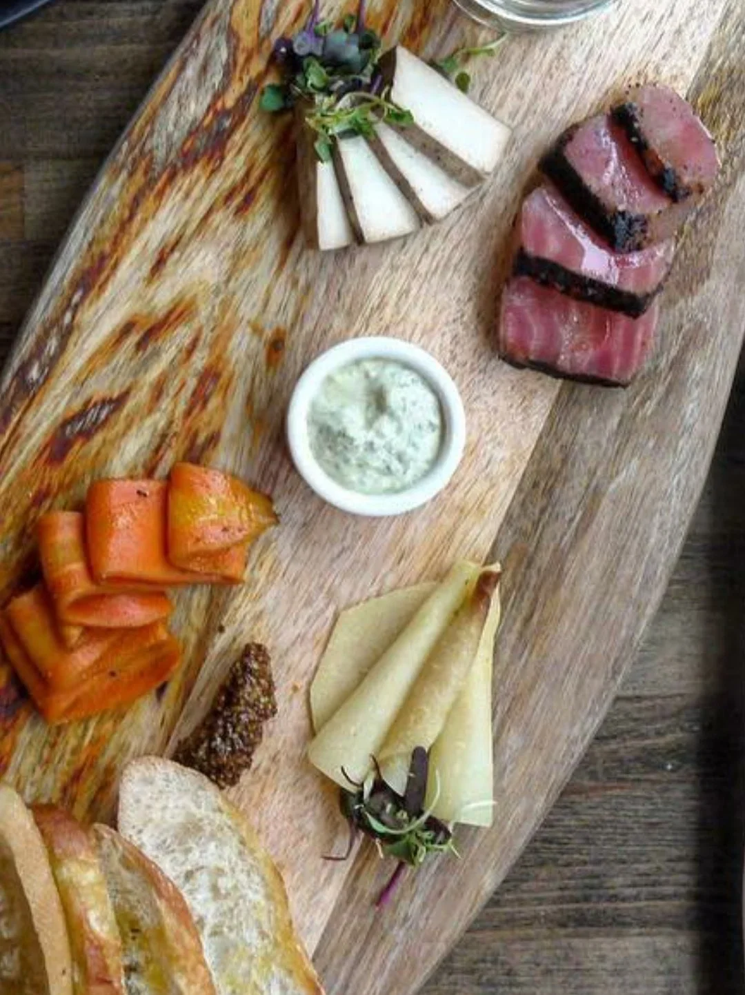 A wooden serving board with cheese slices, smoked salmon, sesame seed-covered bread, a small bowl of herb sauce, and rolled crepes garnished with greens.