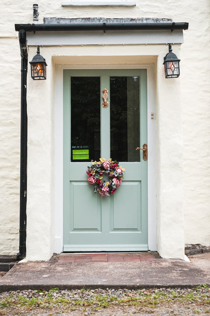A light green front door with a wreath decorated with pink and white flowers and greenery. Two black outdoor lanterns are mounted on either side of the door, and a small step leads up to it. The door is set into a white stucco wall.