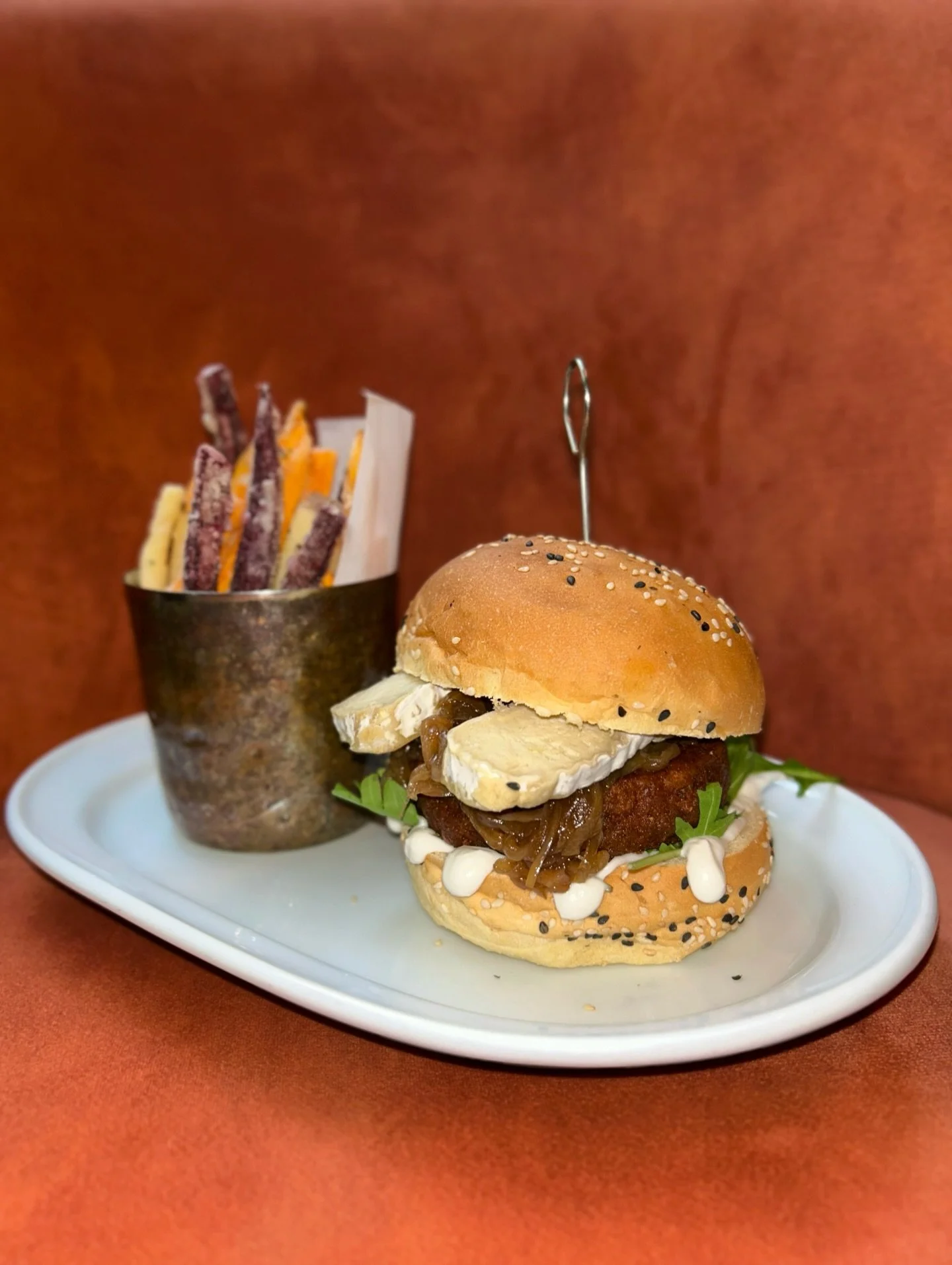 A cheeseburger with pickles, caramelized onions, and mayonnaise on a sesame seed bun, served on a white plate with a side of multicolored snack sticks in a metal cup, against an orange background.
