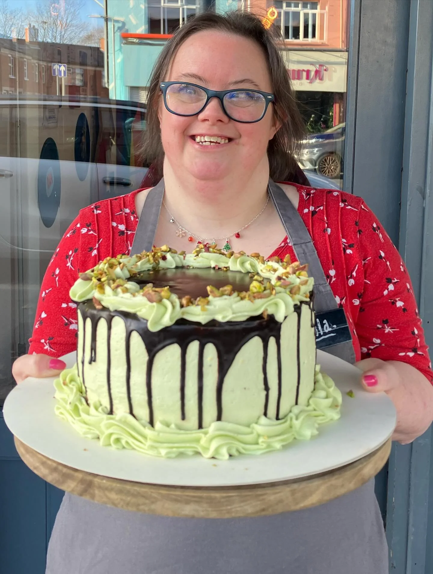 A woman with glasses and a red dress holding a decorated birthday cake outside a storefront.