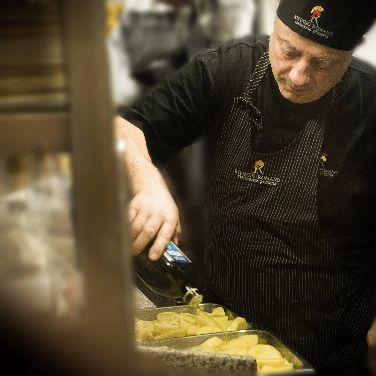 A chef in a black uniform and hat labeled 'Rifugio Romano' is preparing food in a kitchen with yellow potatoes on a tray.