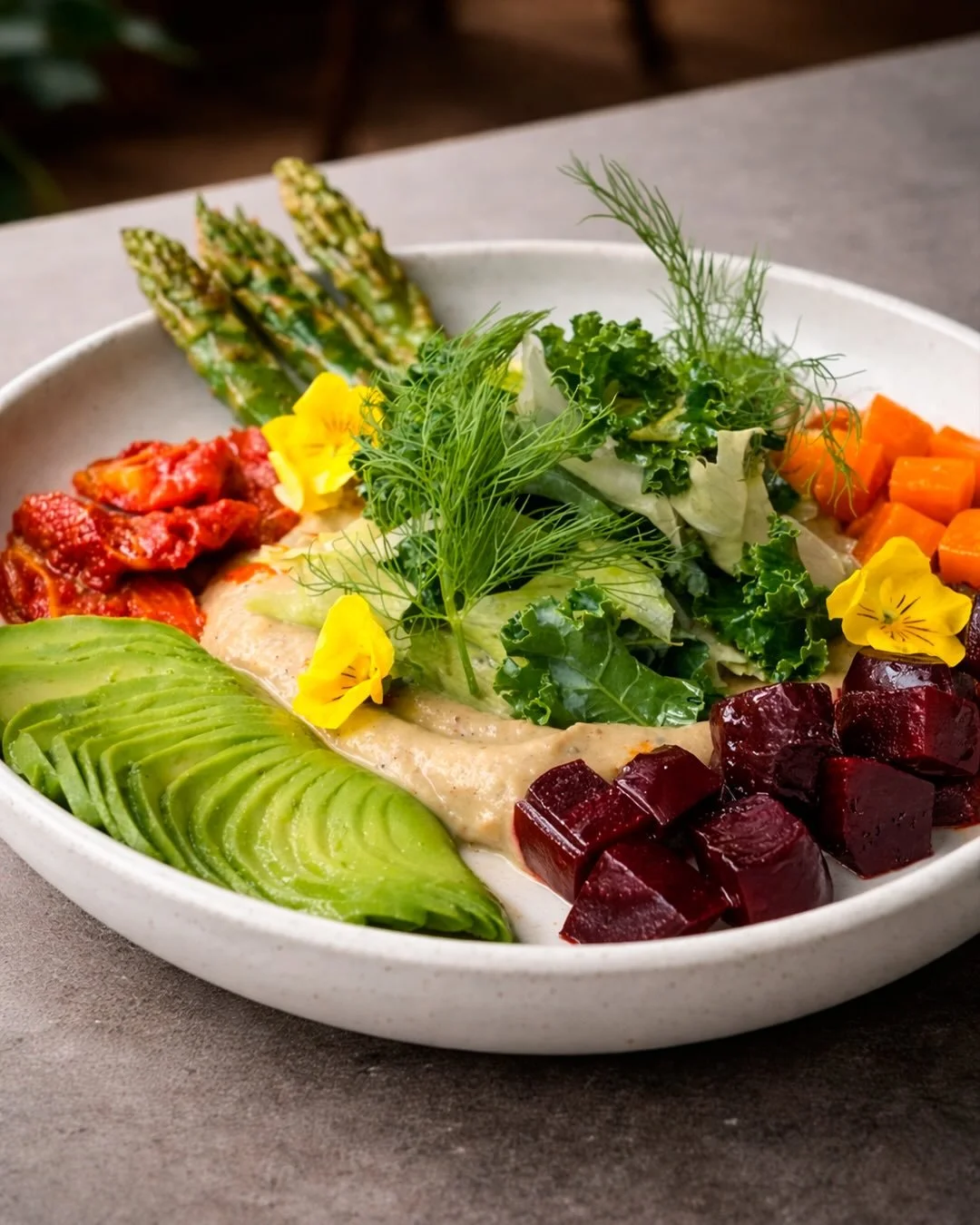 A colorful bowl of assorted vegetables including avocado slices, beet cubes, chopped carrots, leafy greens, asparagus, tomato chunks, and edible flowers, with a creamy hummus or dip in the center.