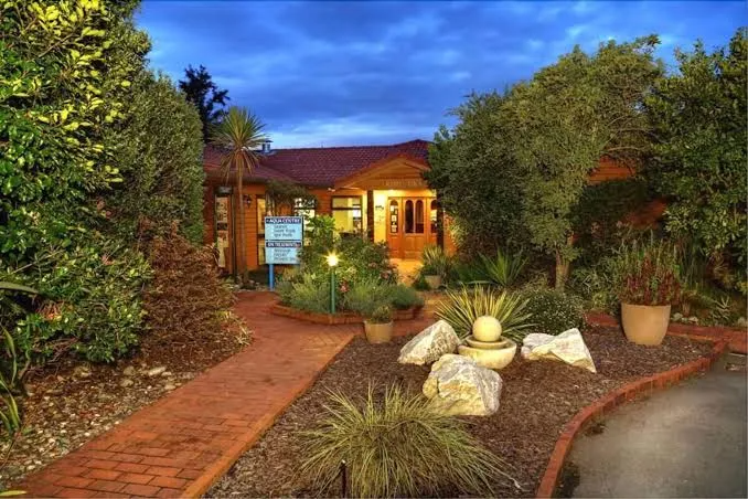 A front yard garden with a brick pathway leading to a house, featuring various plants, rocks, and a decorative fountain, during early evening with lights on.
