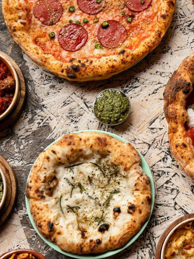 Assorted pizzas with toppings like pepperoni, green peas, and herbs on a rustic wooden table.