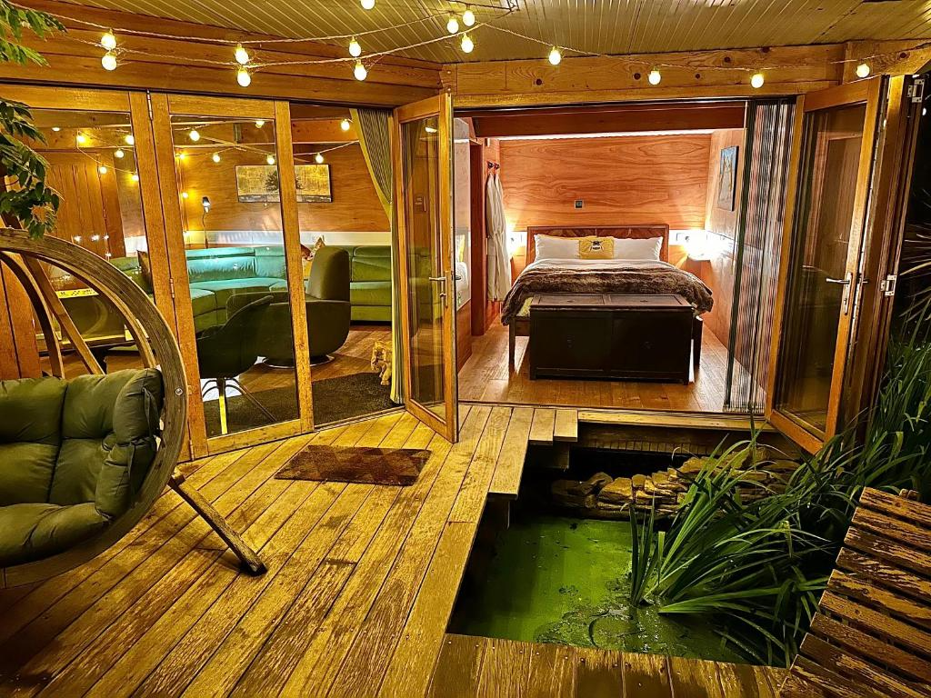 Interior of a cozy wooden cabin with a bedroom, sitting area, and a small pond with plants on a wooden deck, illuminated by string lights.