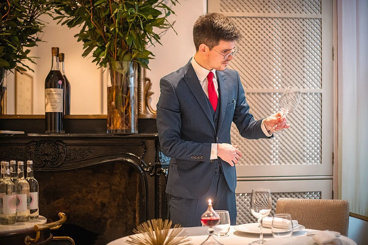 A man in a dark suit, red tie, and glasses inspecting a wine glass in a dining room.
