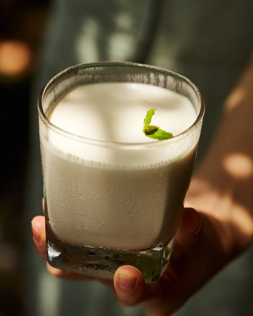 A person holding a glass of a white, creamy beverage garnished with a small green mint leaf.