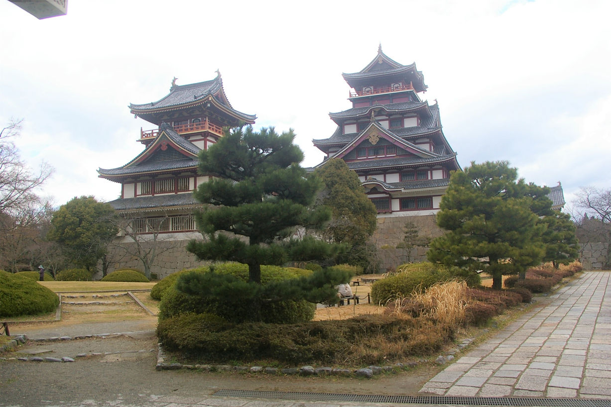 Traditional Japanese castle with multiple tiers and ornate roofs, surrounded by pine trees and a garden with bushes and pathways.