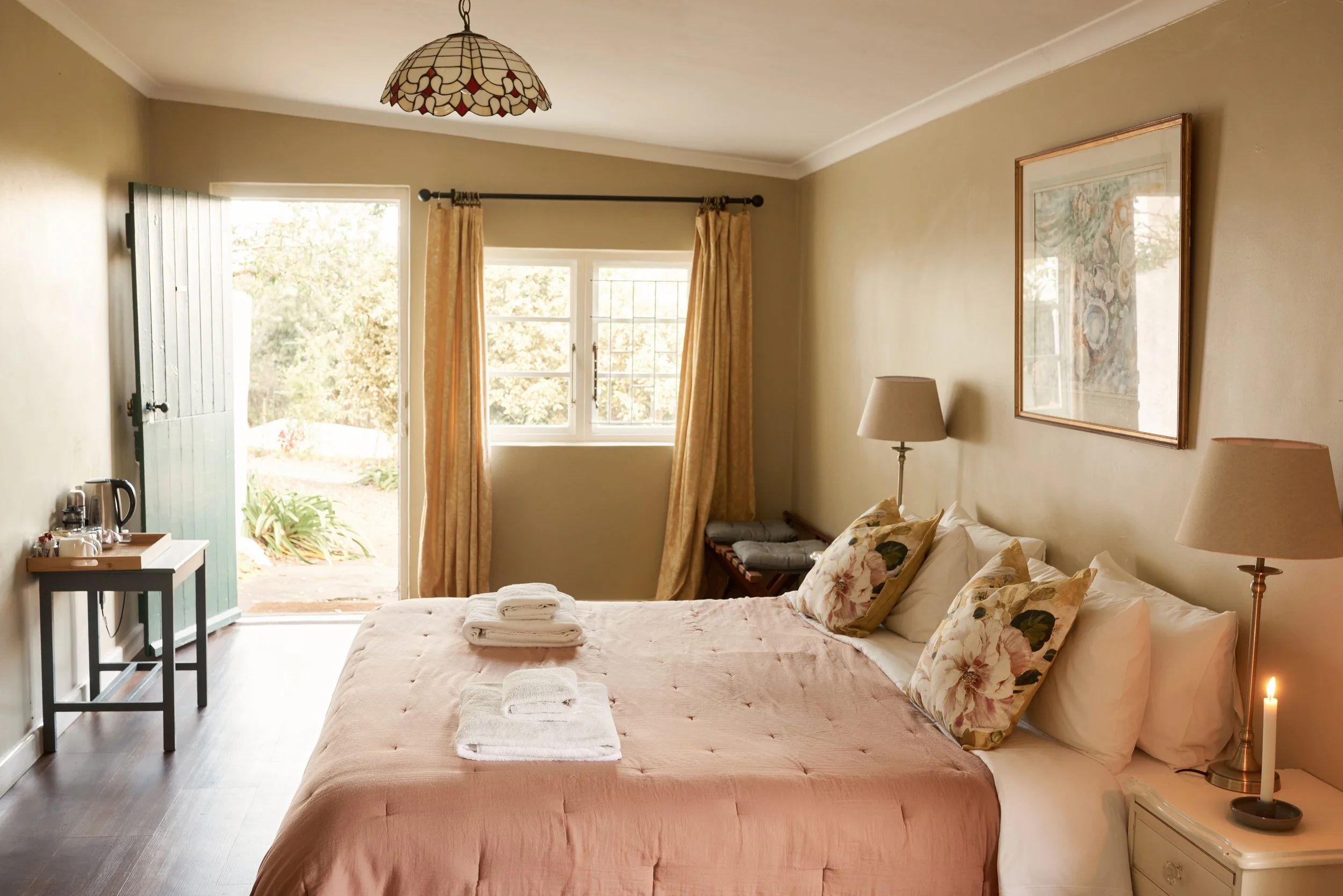 A cozy bedroom with a large bed topped with a pink bedspread and floral pillows. Towels are neatly placed at the foot of the bed. To the right, there is a side table with a lit candle and a lamp. The room has cream-colored walls, a window with curtains, and a framed picture hanging on the wall. An open door reveals a garden outside.