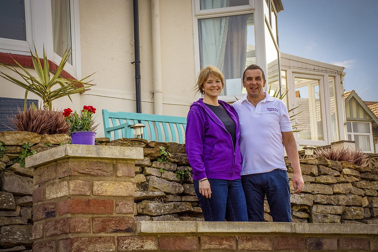 A woman in a purple jacket and a man in a white polo shirt standing on a brick and stone wall outside a house with large windows and a conservatory, smiling at the camera.