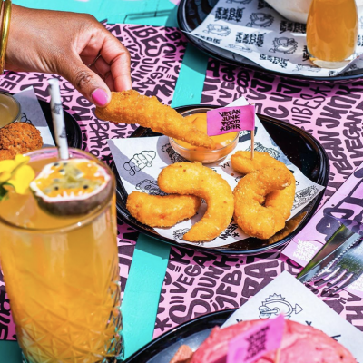 Person dipping a breaded fried onion ring into a small bowl of dipping sauce at a colorful dining table with food and drinks