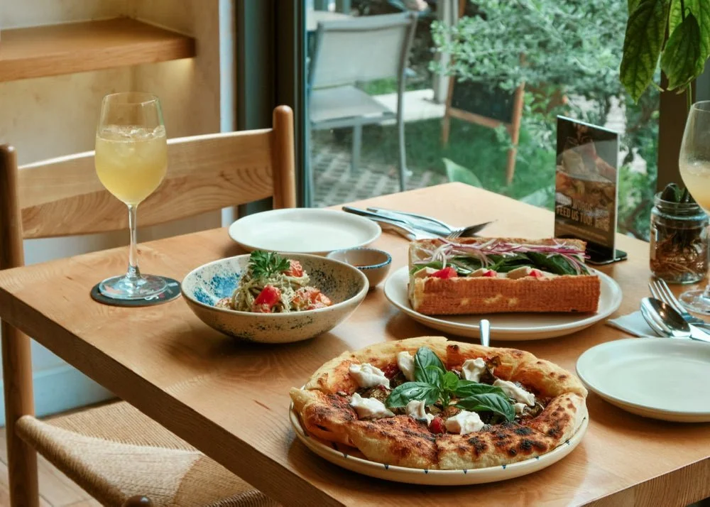 A wooden dining table set with a margarita pizza topped with basil, a bowl of pasta garnished with herbs, a slice of cake, two glasses of white wine or champagne, and empty white plates and utensils, with a window in the background showing greenery outside.
