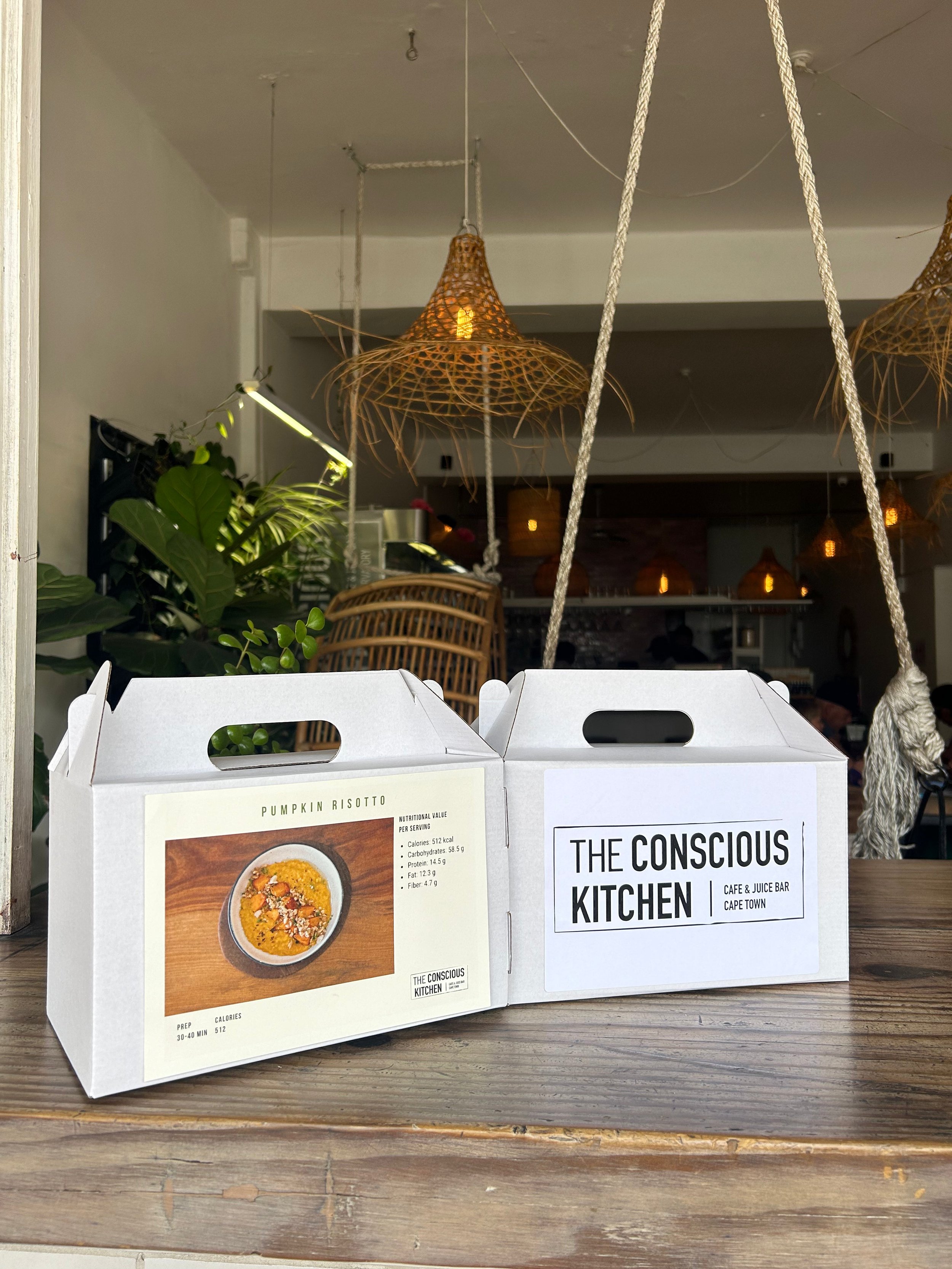 Two white takeout boxes from The Conscious Kitchen restaurant placed on a wooden table. The box on the left shows a pumpkin risotto dish with nutritional information. The box on the right has the restaurant's name and location in Cape Town.
