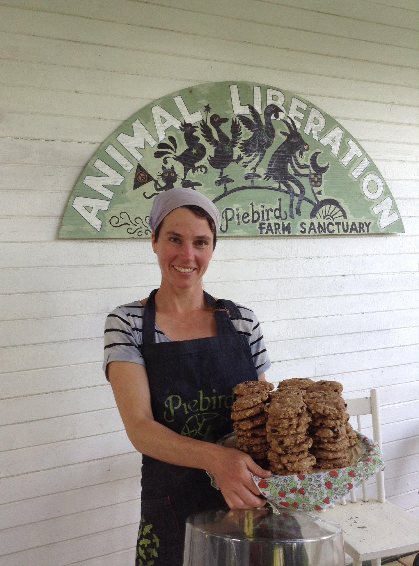 A woman smiling and holding a tray of cookies at a farm sanctuary, standing in front of a sign that reads 'Animal Liberation Piebird Farm Sanctuary' with animal illustrations.
