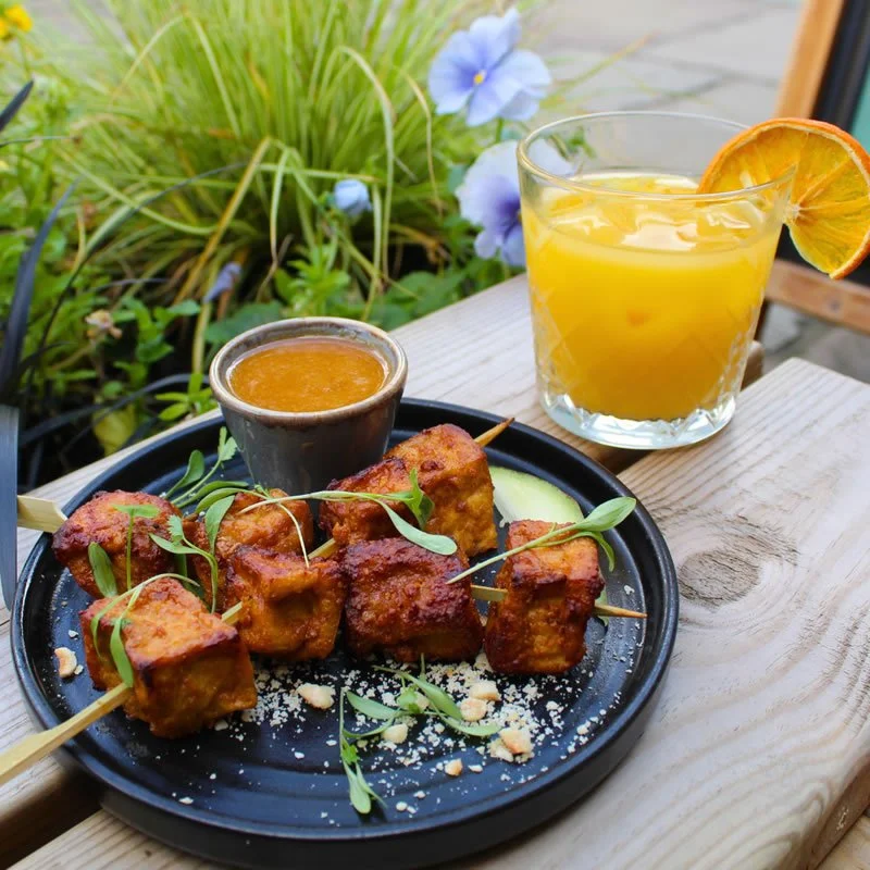 Skewers of grilled chicken with herbs, served with dipping sauce, on a black plate with avocado slices, garnished with microgreens, alongside a glass of orange juice with an orange slice, on a wooden table with greenery and purple flowers in the back