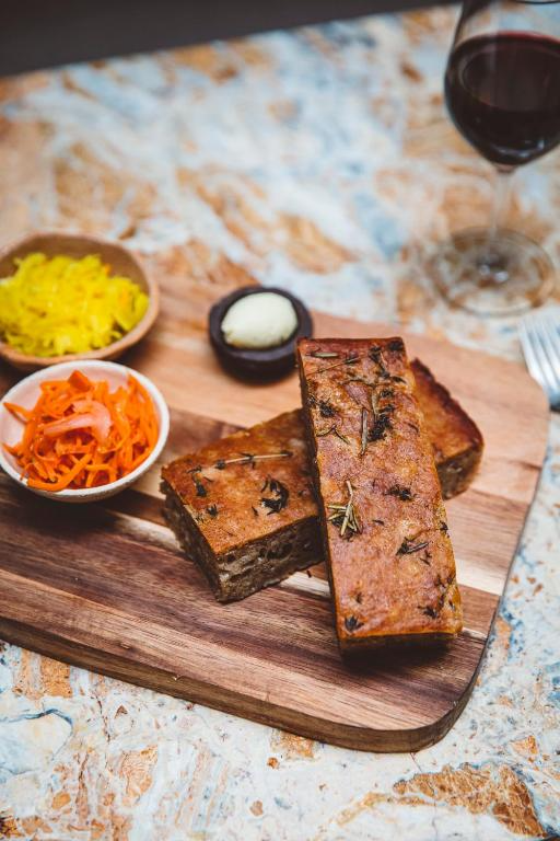 Three slices of meatloaf with herbs on a wooden cutting board, served with yellow and orange shredded vegetables, a small bowl of butter, and a glass of red wine.