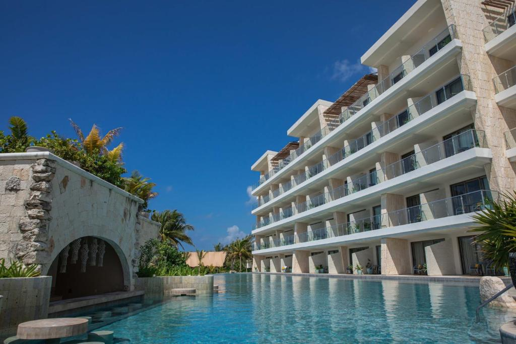 View of a modern hotel with balcony rooms overlooking a swimming pool, blue sky, and palm trees.