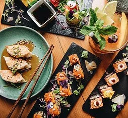 Assorted sushi rolls on a wooden board with garnishes, a plate with sliced sushi, chopsticks, soy sauce, and fresh herbs.