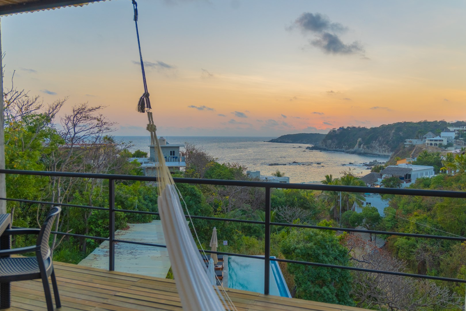 Balcony view overlooking a coastal neighborhood with greenery, houses, and a body of water at sunset, with a hammock hanging from the balcony ceiling.