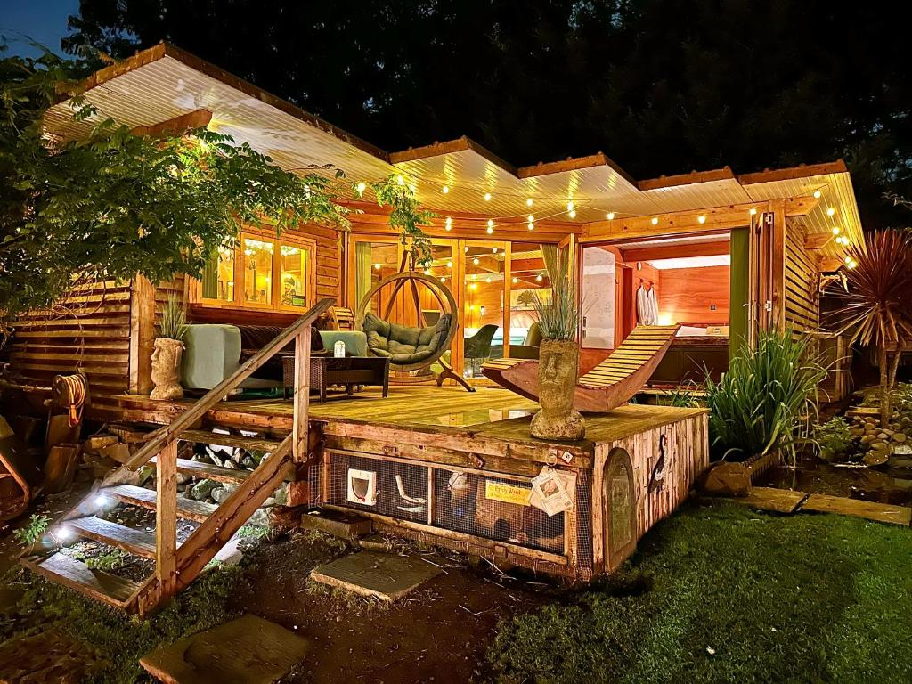 A cozy wooden cabin with a lit outdoor porch, furnished with chairs, a hammock, and string lights, surrounded by greenery at night.