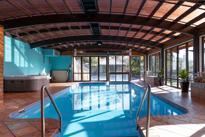 Indoor swimming pool area with large windows, wooden ceiling, lounge chairs, and potted plants
