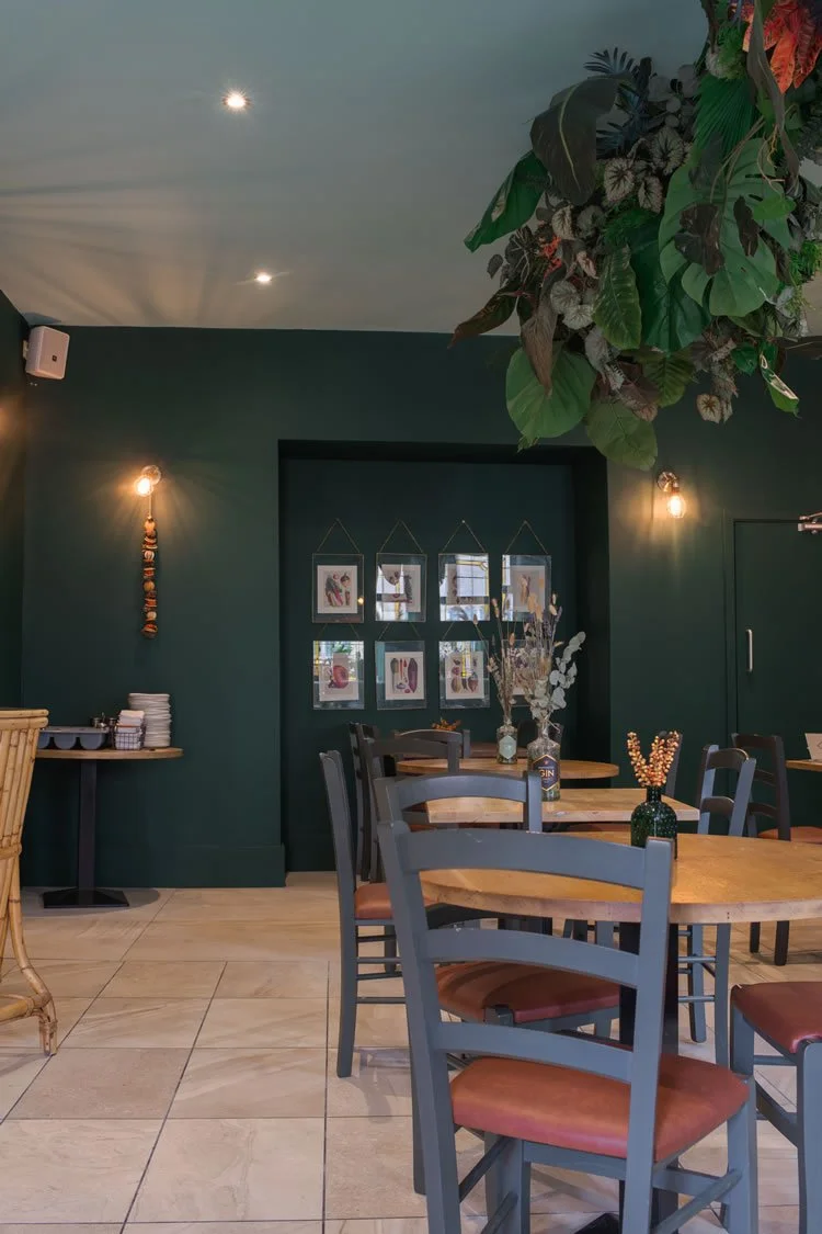 Interior of a restaurant with dark green walls, wooden tables and chairs, and decorative vases with dried plants. Wall art displays framed pictures, and hanging plants are visible from the ceiling.