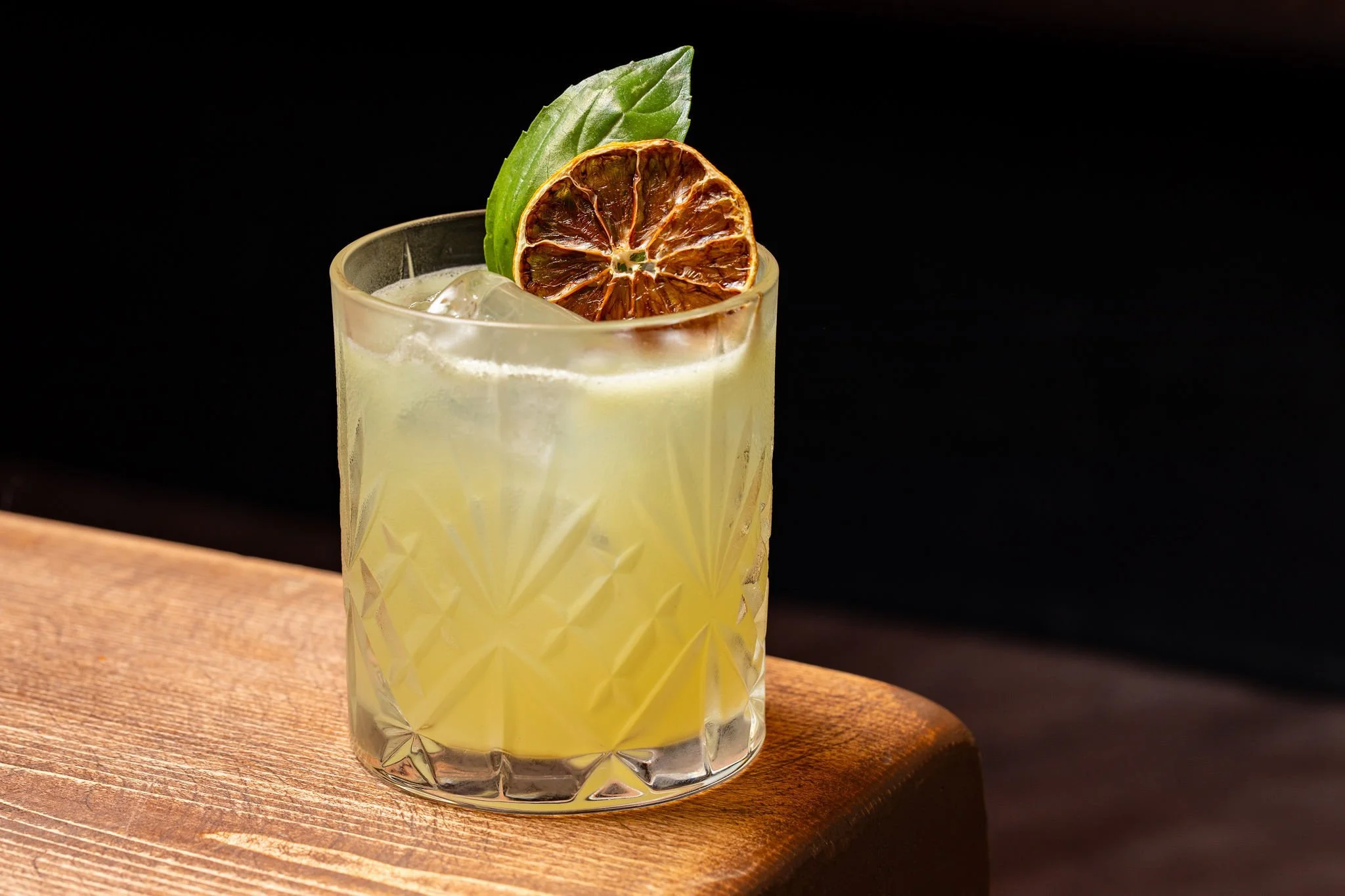 A glass of lemonade garnished with a dried lime slice and a fresh basil leaf on a wooden surface against a dark background.