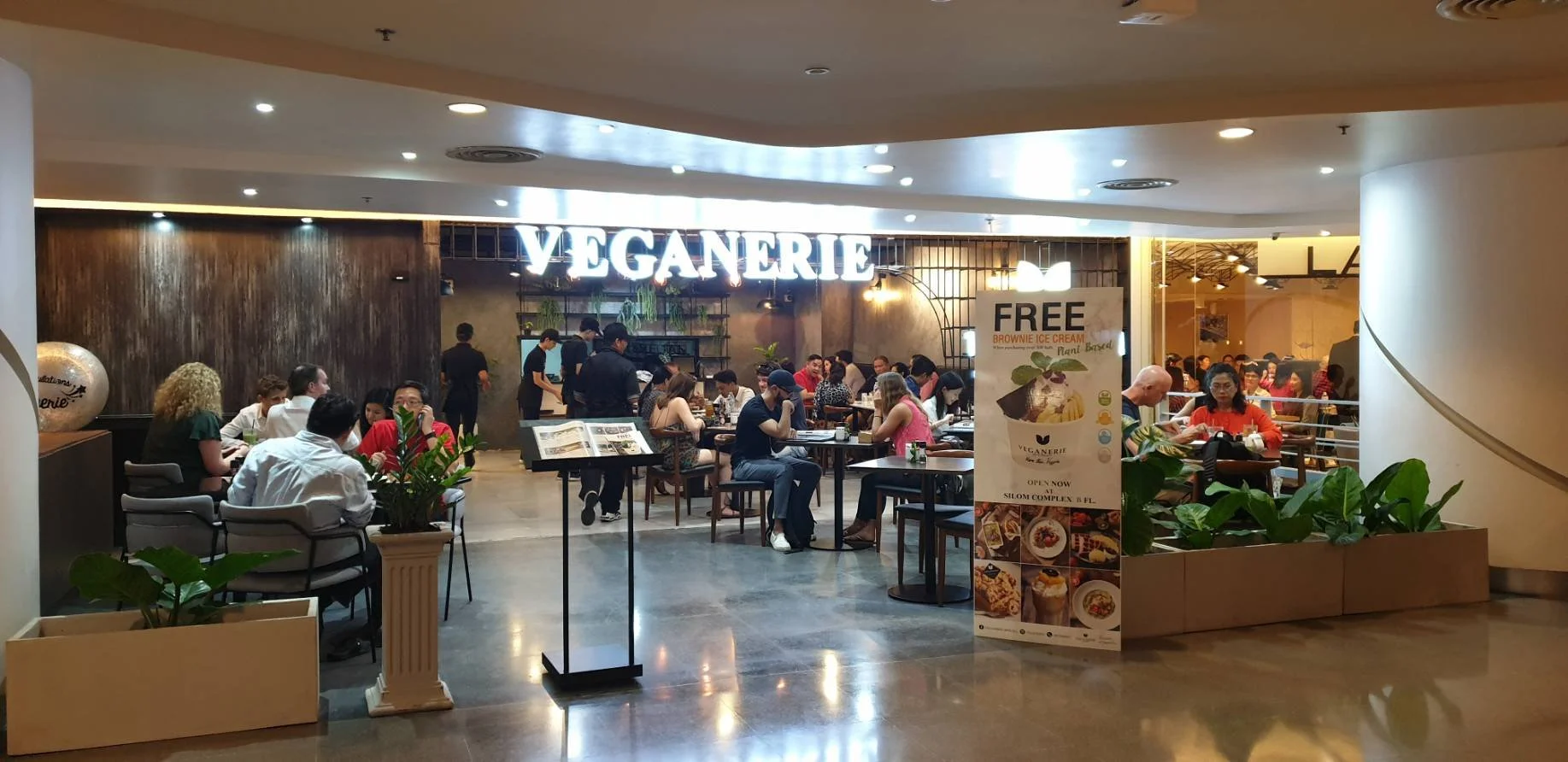 Inside a vegan restaurant, customers are seated and dining, with a large illuminated sign reading 'VEGANERIE' above the entrance, potted plants, and a promotional stand offering free brownie ice cream.
