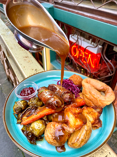 Plate of fried foods, vegetables, and purple cabbage with gravy being poured over it, set on an outdoor table with an 'OPEN' neon sign in the background.