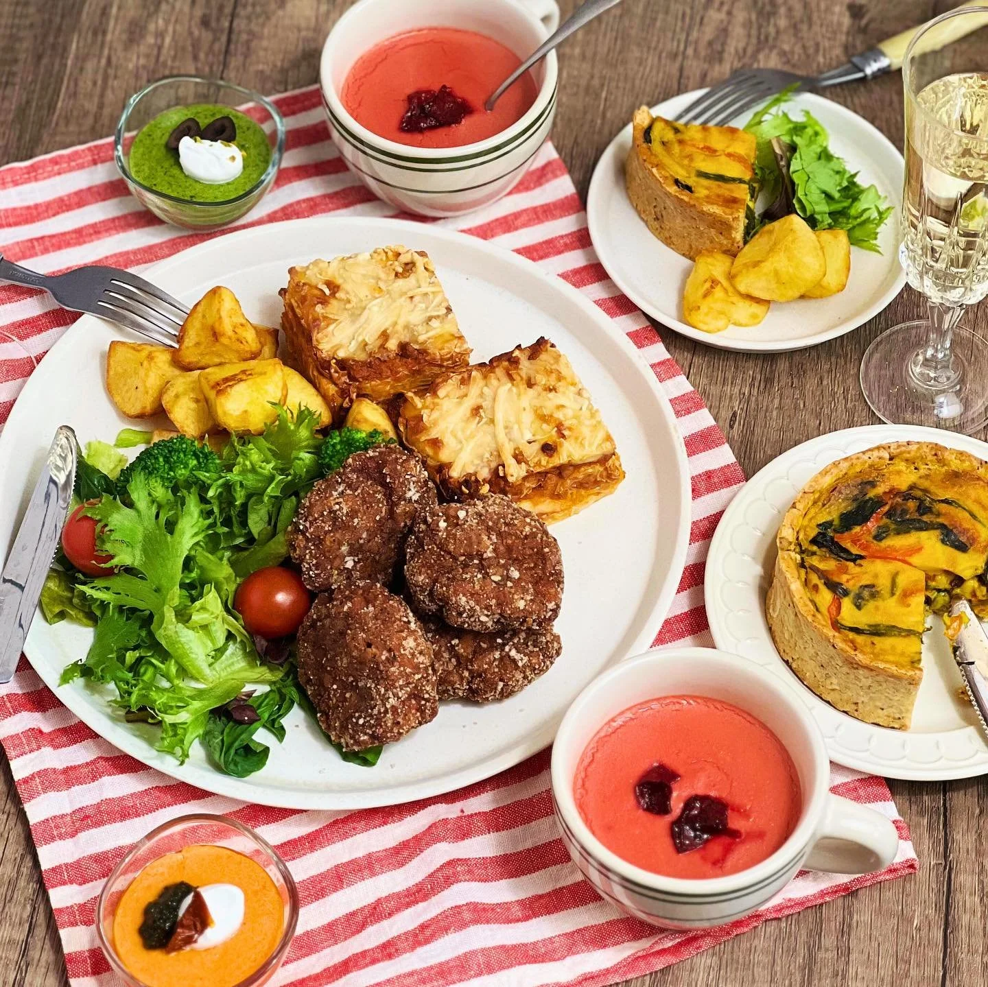 A meal with a large white plate of salad, fried chicken pieces, and stuffed lasagna, with side dishes of sliced vegetable quiche, small salads, and fruit-based soups or desserts on a red and white striped tablecloth.