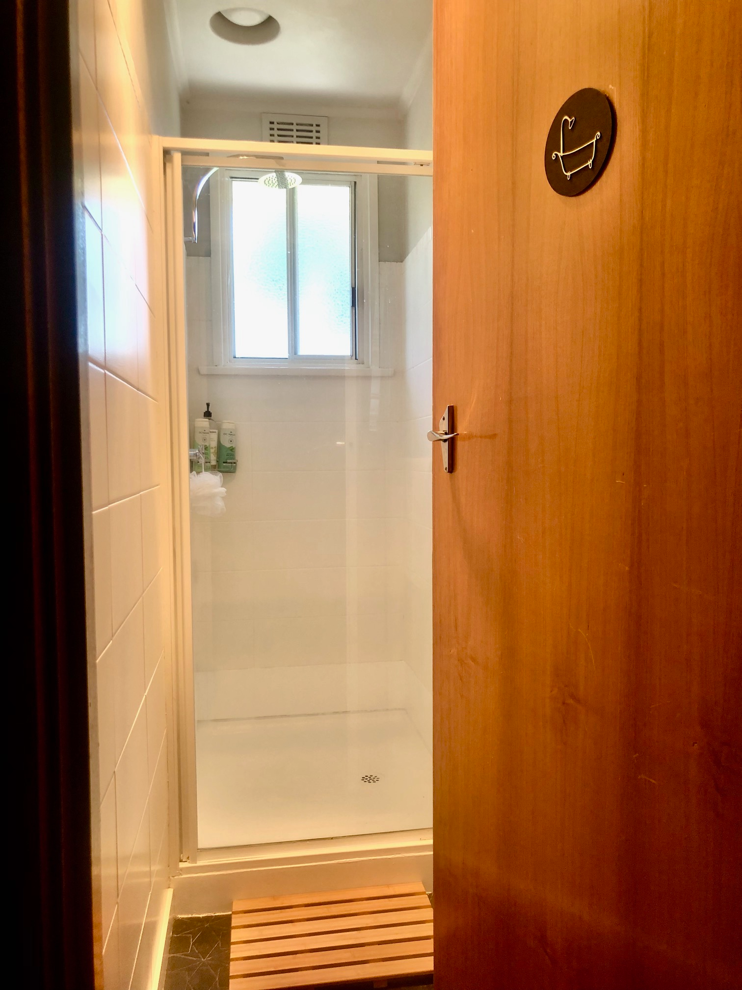 View of a bathroom shower with a frosted window, toiletries, and a wooden bench next to a wooden surface in the foreground.