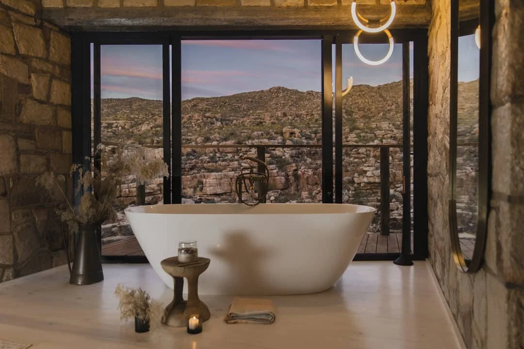 Modern bathroom with a freestanding bathtub, large front windows with a mountain view, and decorative lighting fixtures.