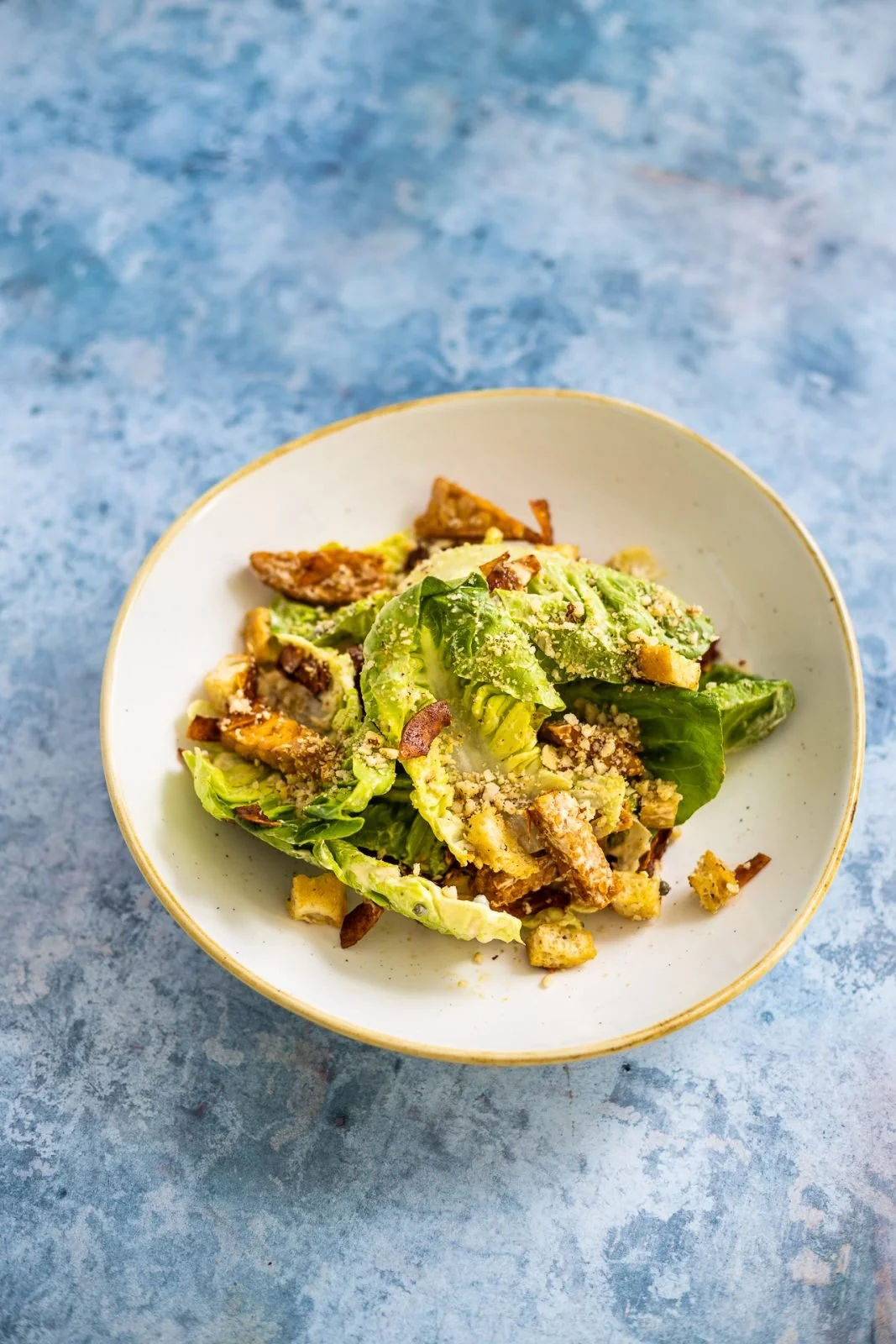 A bowl of Caesar salad with romaine lettuce, croutons, and grated cheese on a blue textured surface.