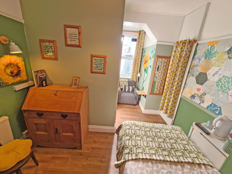 A cozy bedroom with green walls, a bed with a geometric blanket, a white side table with a lamp and kettle, and a large colorful quilt on the wall. There is a chest of drawers with framed photos, and a sitting area with a window in the background.