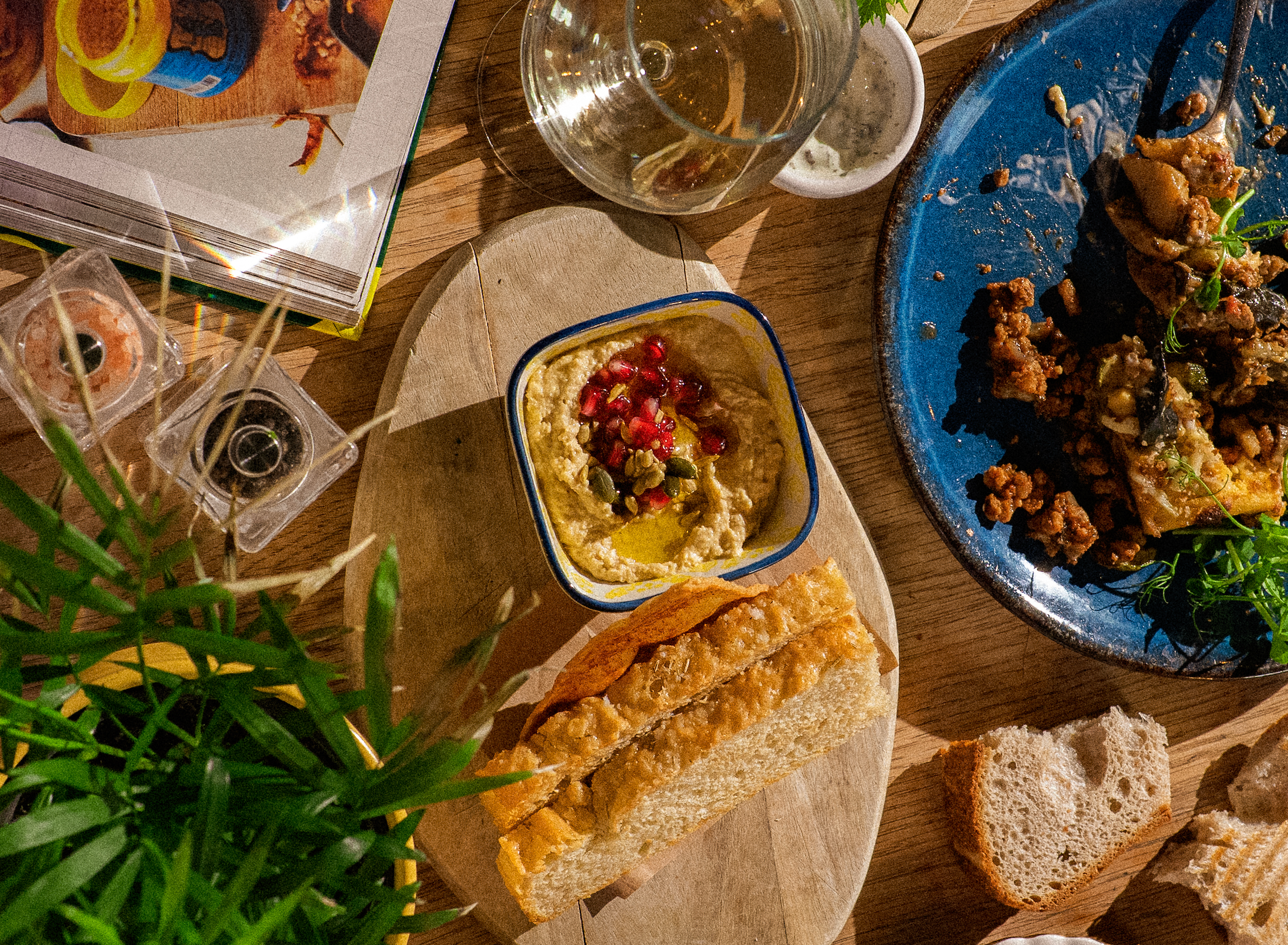 A wooden table with various dishes and drinks, including a glass of white wine, a bowl of hummus topped with pomegranate seeds, slices of bread, and a blue plate with fried food garnished with greens. There are some snacks and a magazine on the table