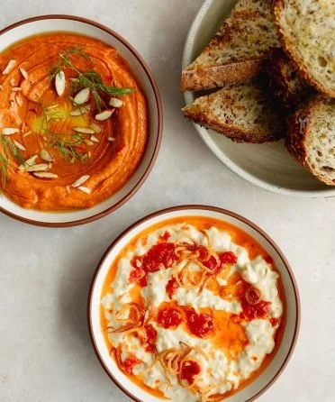 Bowl of red lentil soup garnished with herbs and sunflower seeds, bowl of creamy ricotta cheese topped with tomato sauce, and a plate of toasted bread slices.