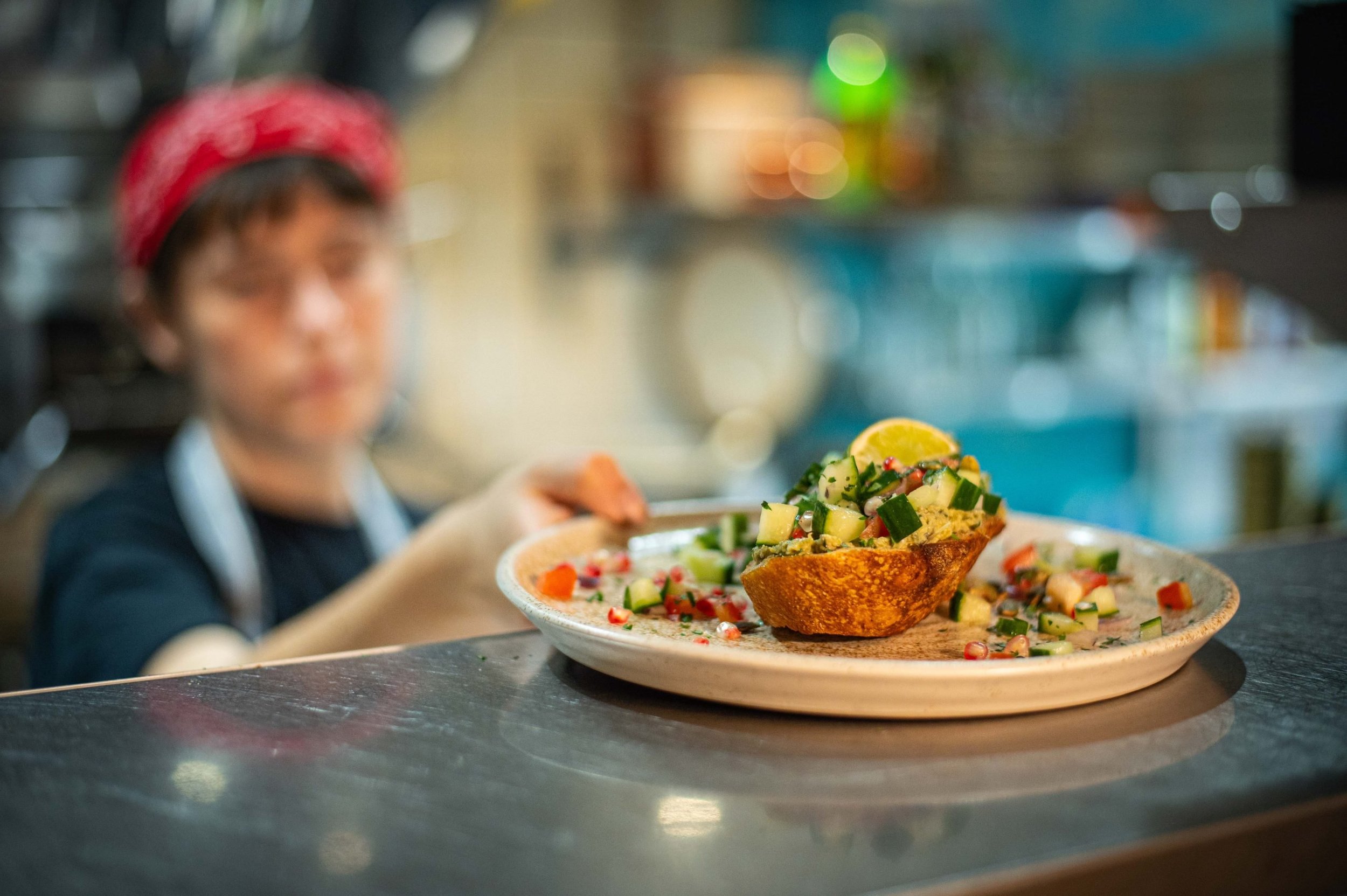 A person with a red bandana on their head serving a colorful salad with diced vegetables and lemon on a plate in a kitchen setting.