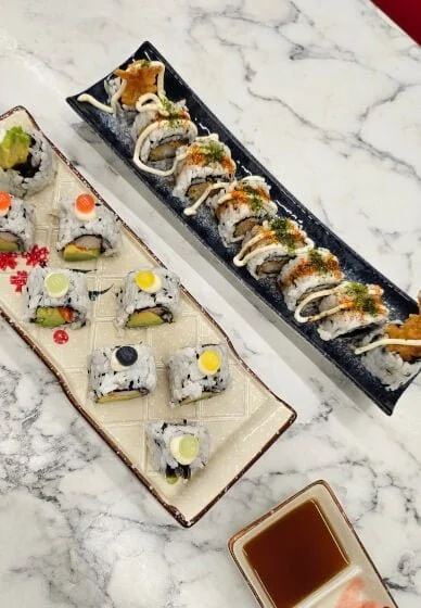 Various sushi rolls on two rectangular plates, with a small bowl of soy sauce nearby, on a marble surface.