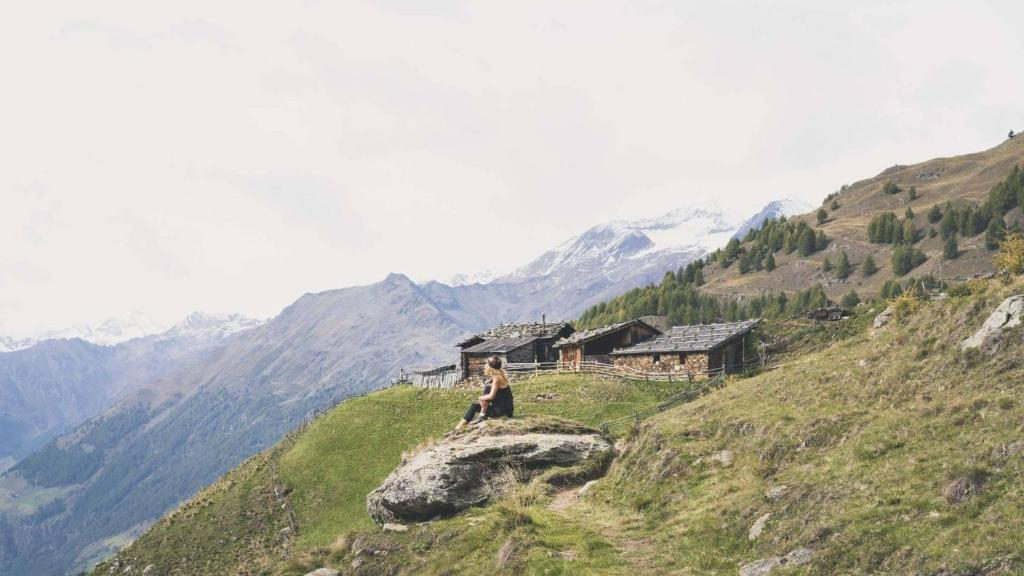 A woman sitting on a large rock on a grassy hillside with mountains and a few rustic wooden buildings in the background.