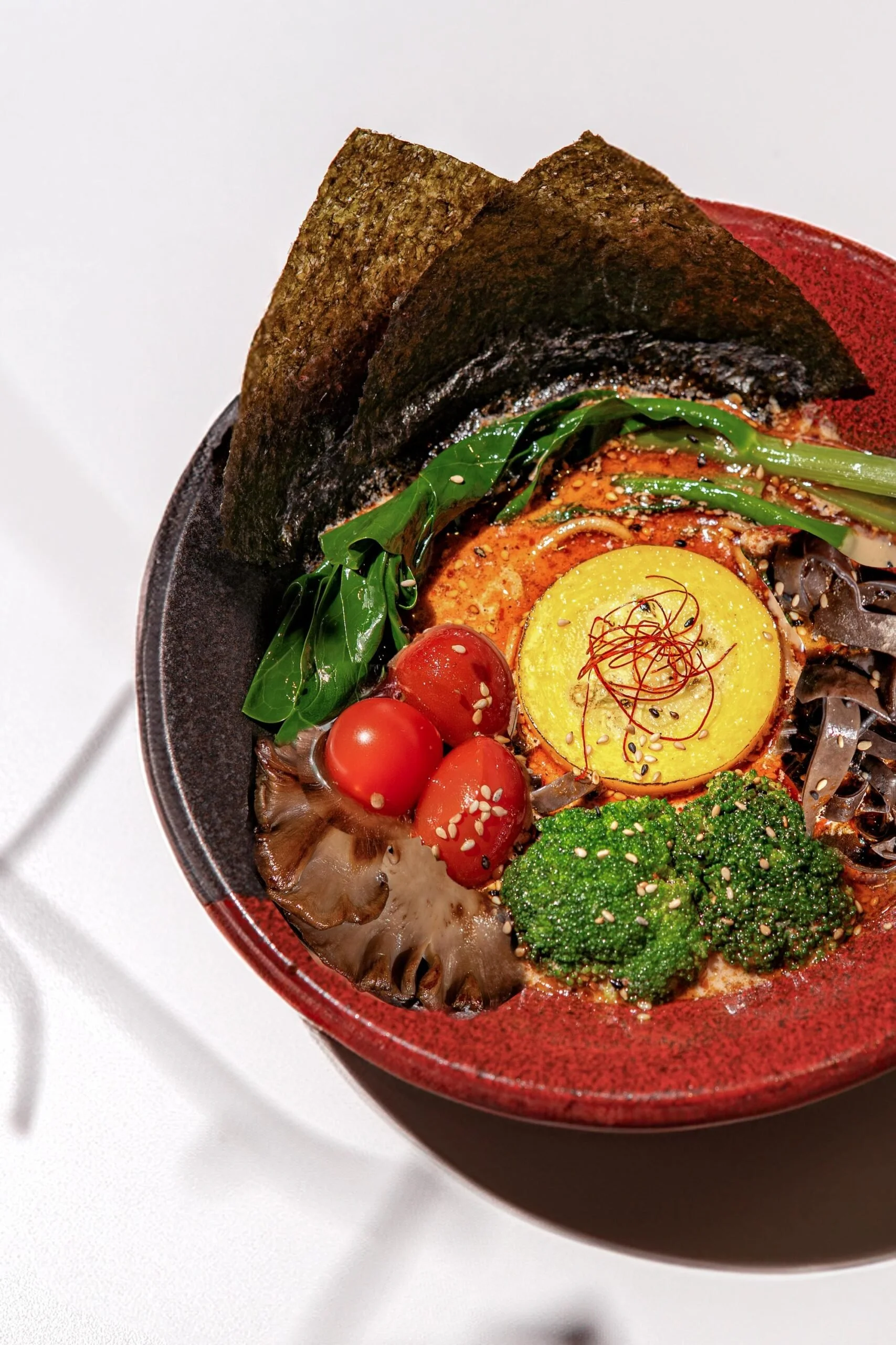 Close-up of a bowl of ramen noodles topped with seaweed, cherry tomatoes, broccoli, a boiled egg, and green onions.