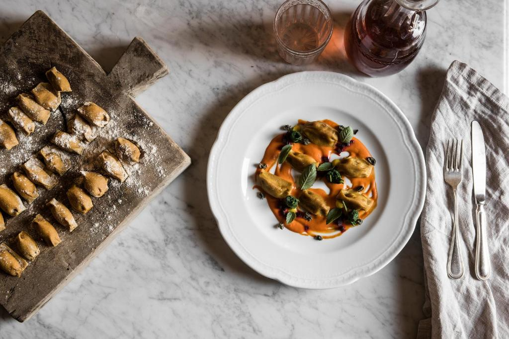 A plate of Italian pasta with ravioli topped with basil and black olives, served with a red sauce, on a white decorative plate. To the left, there is a wooden cutting board with uncooked ravioli dusted with flour. A glass of rosé wine and a carafe are at the top, with a fork and knife on a white napkin to the right.