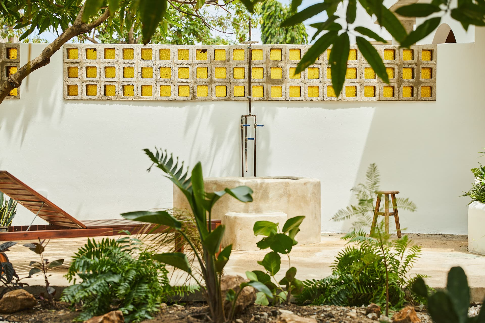 Garden patio with surrounding greenery, a white wall with yellow brick accents, a stone water feature, a wooden chair, and plants in the foreground.