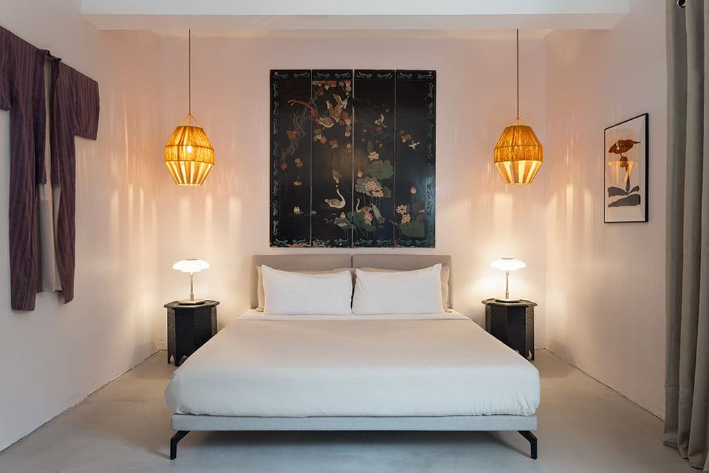 A bedroom with a bed, two nightstands with lamps, and wall art featuring traditional Asian artwork, in a softly lit room with purple curtains and beige walls.