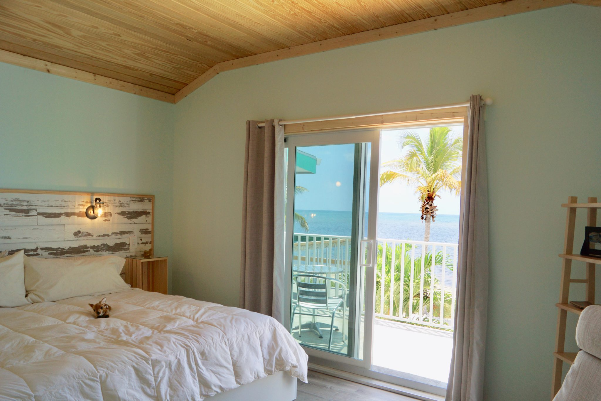 A bedroom with a sliding glass door leading to a balcony overlooking the ocean, with a palm tree visible outside. The room has a wooden ceiling, a white bed with a small dog or puppy lying on it, and beige curtains on the door.