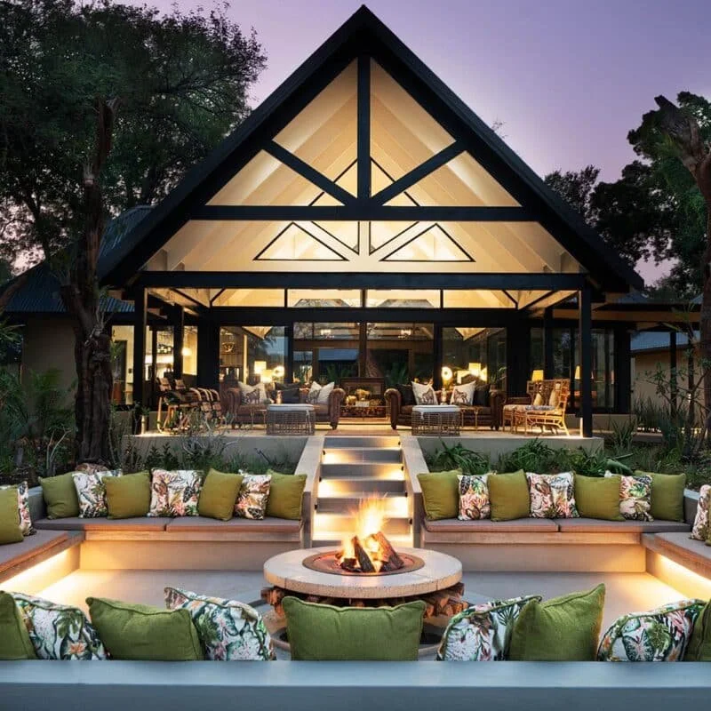 Exterior view of a modern house with an open living area, large glass windows, furnished outdoor seating with green and floral pillows around a fire pit, and lush trees in the background during evening.