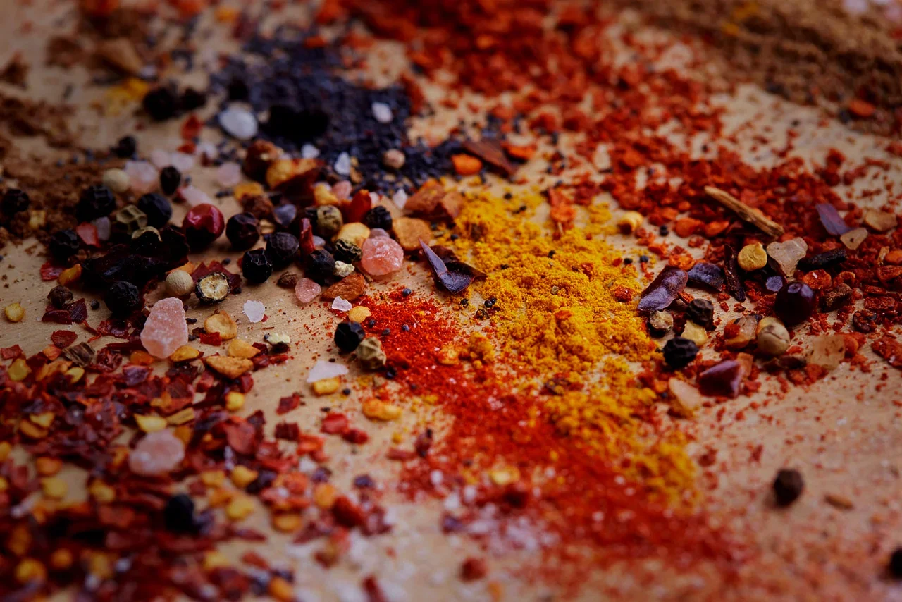 Various colorful spices scattered on a flat surface, including peppercorns, pink Himalayan salt, turmeric, and chili powder.